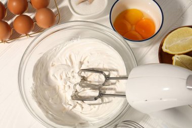Beyaz ahşap masada çırpılmış krema, yumurta ve diğer ürünlerle el çırpma makinesi.