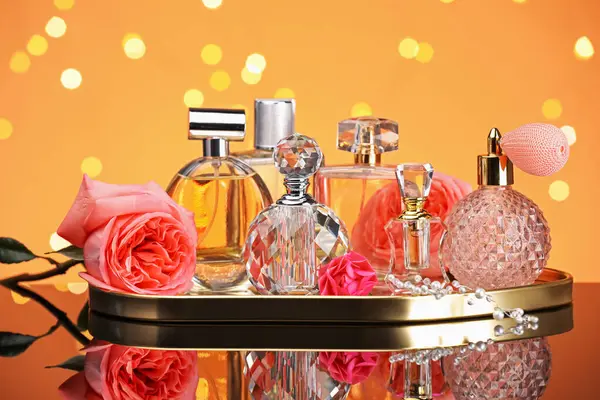 Farklı parfüm şişeleri ve çiçekler ayna yüzeyinde turuncu arka planda bulanık ışıklarla, yakın plan