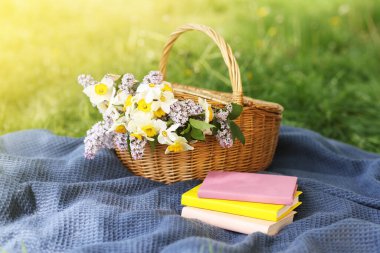 Yığınla kitap, battaniye ve hasır sepet. Yeşil çimlerin üzerinde çiçekler.