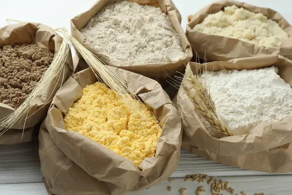 Beyaz ahşap masadaki kağıt torbalarda farklı türde un, yakın plan.