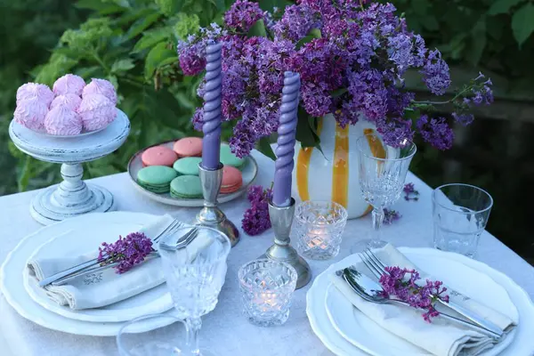 Güzel leylak çiçekleri, tatlılar, tabaklar, çatal bıçak takımı, bardaklar ve mumlar bahçedeki masada.