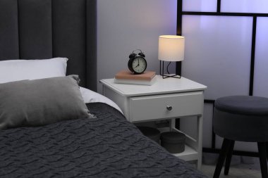 İçinde lamba, çalar saat ve yatağın yanında kitaplar olan komodin.