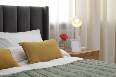 Yatağın yanında lamba, çiçek ve çerçeve olan komodin.