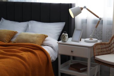 Yatağın yanında lamba, çerçeve ve alarmlı komodin.