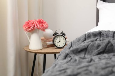 Yatağın yanında çalar saati, çiçekleri ve kitapları olan bir komodin.