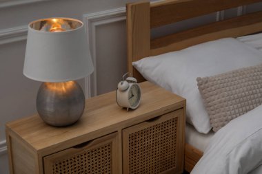 Yatağın yanında lamba ve çalar saat olan komodinin yanında.