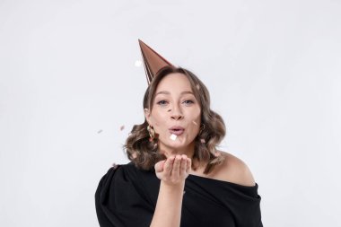 Doğum günü kutlaması. Parti konisi olan bir kadın beyaz arka planda konfeti üflüyor.