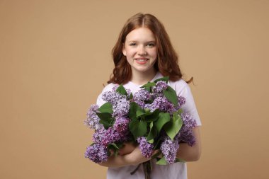 Bej arka planda bir demet leylak çiçeğiyle gülümseyen genç kız.