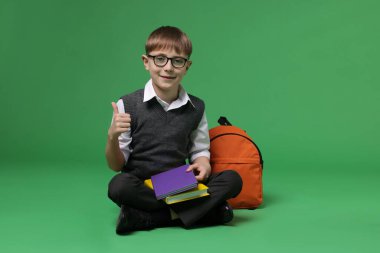 Sırt çantalı ve kitaplı mutlu okul çocuğu yeşil arka planda başparmak gösteriyor, mesaj için yer var.