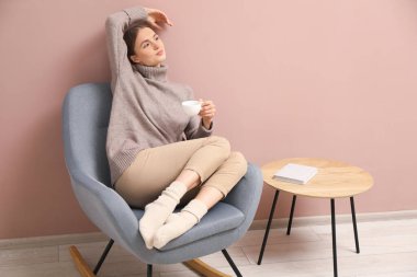 Güzel bir kadın, bir bardak içkiyle bej duvarın yanındaki sallanan sandalyede dinleniyor.