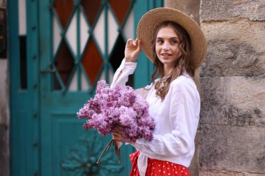 Şapkalı gülümseyen kadın, bina yakınlarında bir demet leylak çiçeğiyle.