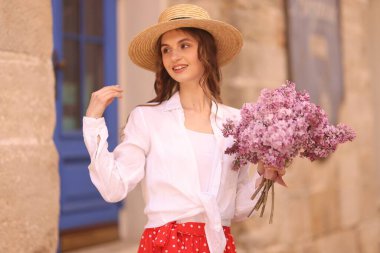 Şapkalı gülümseyen kadın, bina yakınlarında bir demet leylak çiçeğiyle.