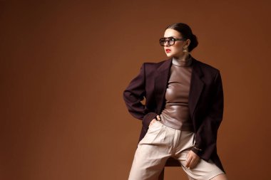 Kahverengi arka planda sandalyede şık giyinmiş genç bir kadın, mesaj için yer var.