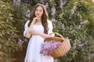 Güzel bir kadın, çalılıkların yanında bir sepet leylak çiçeği taşıyor.