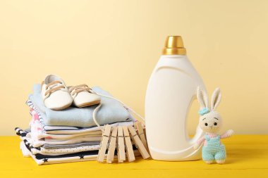 Kumaş yumuşatıcısı, bebek kıyafetleri, mandallar ve bej arka planda sarı ahşap masada oyuncak.