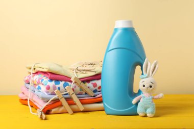 Kumaş yumuşatıcısı, bebek kıyafetleri, mandallar ve bej arka planda sarı ahşap masada oyuncak.