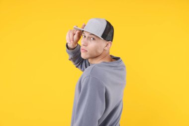 Sarı arka planda şık bir beyzbol şapkası takan genç adam.