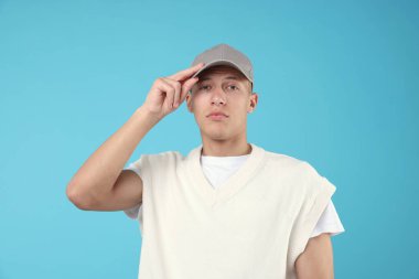 Genç adam açık mavi arka planda şık gri beyzbol şapkası giyiyor.