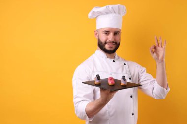 Üniformalı mutlu pastacı elinde eklerlerle turuncu arka planda iyi bir jest yapıyor.