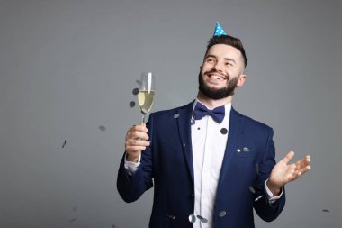 Doğum günün kutlu olsun. Gri arka planda konfeti banyosu altında parti şapkası ve bir bardak şampanyası olan yakışıklı bir adam.
