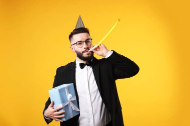 Doğum günün kutlu olsun. Turuncu arka planda parti şapkası, üfleme makinesi ve hediye kutusu olan yakışıklı bir adam.