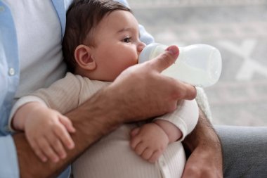 Babası küçük bebeğini evdeki koltukta biberonla besliyor.