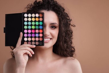 Gülümseyen kadın bej arka planda göz farı paletiyle gözlerini kapatıyor.