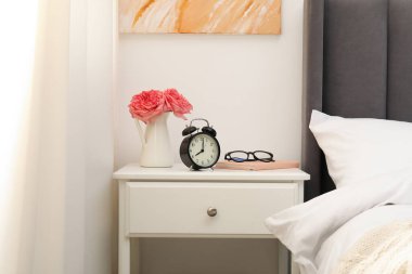 Yatağın yanında çalar saati, çiçekleri ve bardakları olan bir komodin.