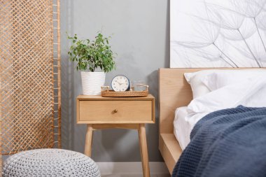 Yatağın yanında ev bitkisi, çalar saat ve yatağın yanında bir bardak su olan komodin.