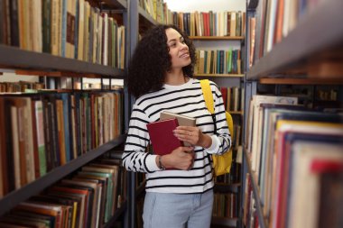 Halk kütüphanesinde rafların yanında kitapları ve sırt çantaları olan bir kadın.