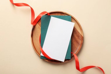Boş davetiye kartı, zarf, kırmızı kurdele ve bej arka planda ahşap tepsi.