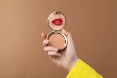 Kırmızı dudaklı güzel bir kadın soluk kahverengi arka planda pudra aynasına bakıyor.