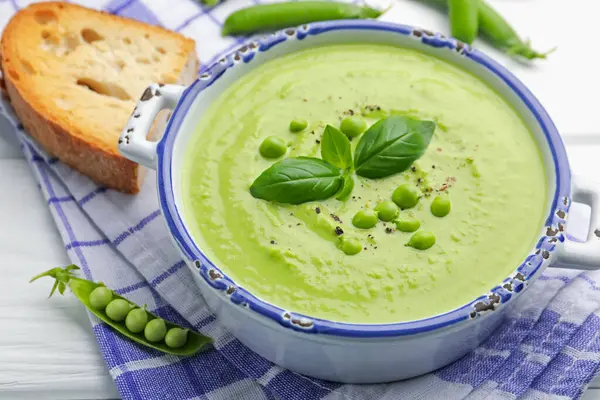 Beyaz ahşap masada servis edilen fesleğenli bezelye kremalı çorba.
