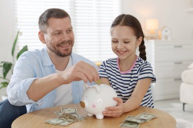 Baba ve kızı kapalı bir masadaki kumbaraya cep harçlığı koyuyor.