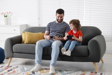 Mutlu baba ve oğlu evdeki kanepede oyun kumandalarıyla oynuyorlar.