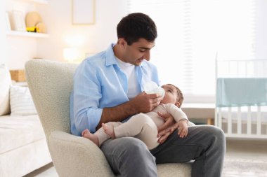 Baba küçük bebeğini evdeki koltukta şişeyle besliyor.