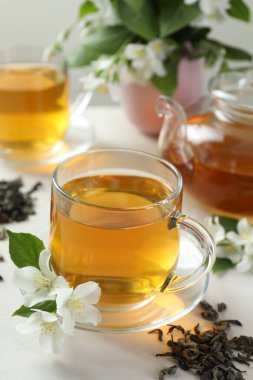 Cam bardaklarda, çaydanlıkta, çiçeklerde ve beyaz masada kurumuş yapraklarda Aromatik yasemin çayı.