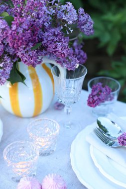 Güzel leylak çiçekleri, tabaklar, çatal bıçak takımı, bardaklar ve yanan mumlar bahçede, yakın plan.