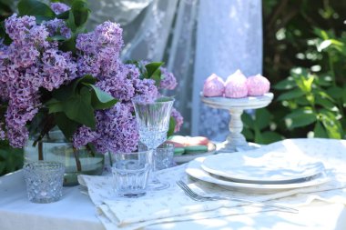 Bahçedeki masada güzel leylak çiçekleri, tatlılar, tabaklar, çatal bıçaklar ve bardaklar.