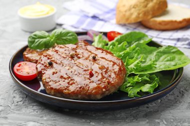 Lezzetli hamburger köfteleri, domates, marul, çörekler ve baharatlar gri masada, yakın plan.
