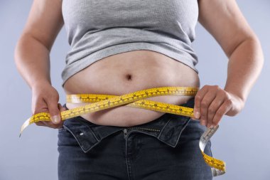 Kilolu bir kadın göbeğini açık arkaplanda bantla ölçüyor.