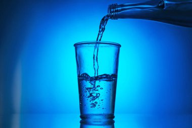 Mavi arkaplanda şişeden cama su dökmek. Arka ışıklandırma