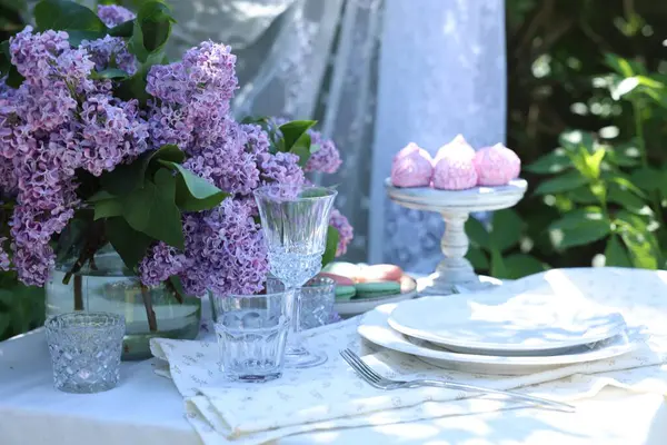 Bahçedeki masada güzel leylak çiçekleri, tatlılar, tabaklar, çatal bıçaklar ve bardaklar.