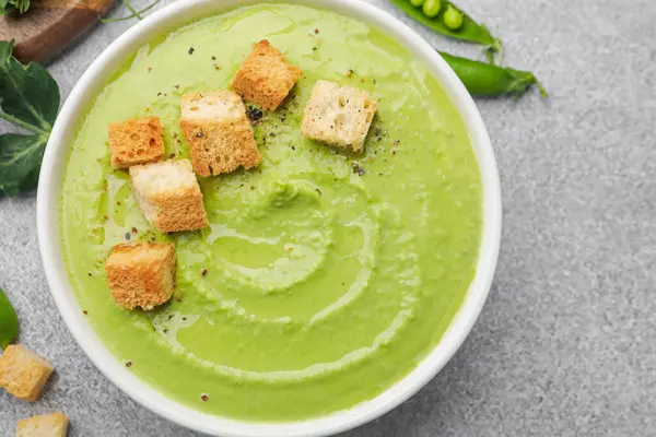Gri masada servis edilen kızarmış ekmekli bezelye kremalı çorba.