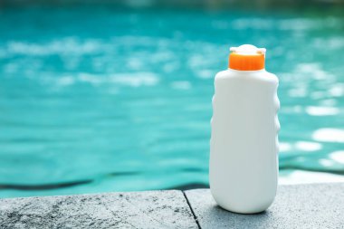 Dışarıdaki yüzme havuzunun yakınındaki taş yüzeyde bir şişe güneş koruması ürünü. Metin için yer var.