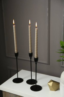 Zarif şamdanlar, yanan mumlar ve gri duvarın yanındaki şifoniyerin üzerindeki diğer dekorlar.