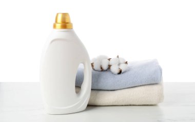 Bir şişe yumuşatıcı, üst üste yığılmış giysiler ve pamuk çiçekleri beyaz arka plana karşı mermer masa