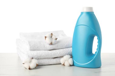 Bir şişe yumuşatıcı, beyaz arkaplana karşı mermer masada istiflenmiş havlular ve pamuk çiçekleri.