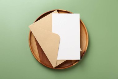 Açık yeşil arka planda boş davetiye, zarf ve ahşap tepsi, üst görünüm