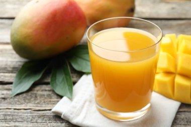 Bardakta lezzetli mango suyu, taze meyve ve ahşap masada yeşil yapraklar, yakın plan.